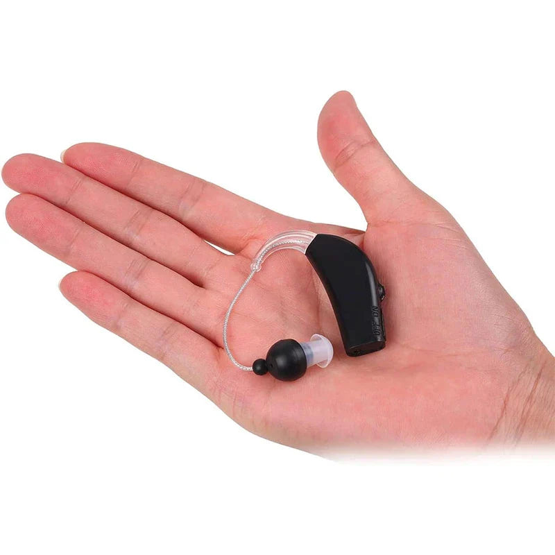 A hand holding a black rechargeable hearing aid with clear ear tube and black ear tip against white background