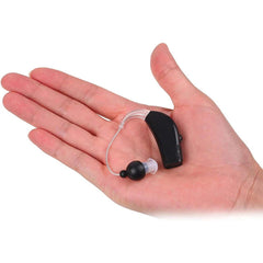 A hand holding a black rechargeable hearing aid with clear ear tube and black ear tip against white background