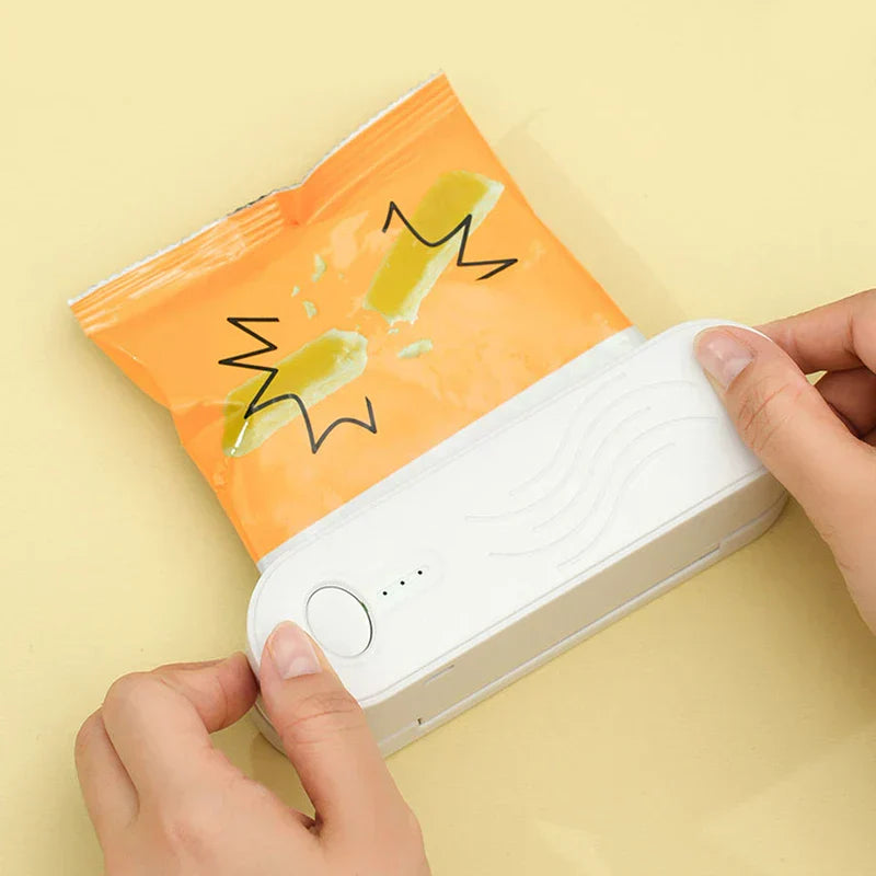 Hands holding a white mini bag heat sealer pressed against the open top of an orange snack bag on a pale yellow surface