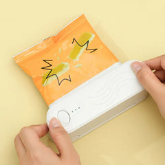 Hands holding a white mini bag heat sealer pressed against the open top of an orange snack bag on a pale yellow surface