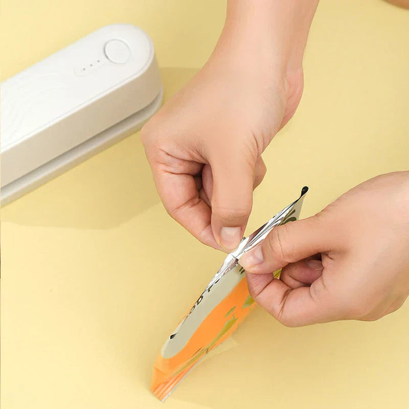 Hands holding a sealed plastic snack bag, with a white mini bag heat sealer resting on a pale yellow surface in the upper left