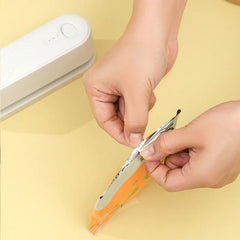 Hands holding a sealed plastic snack bag, with a white mini bag heat sealer resting on a pale yellow surface in the upper left
