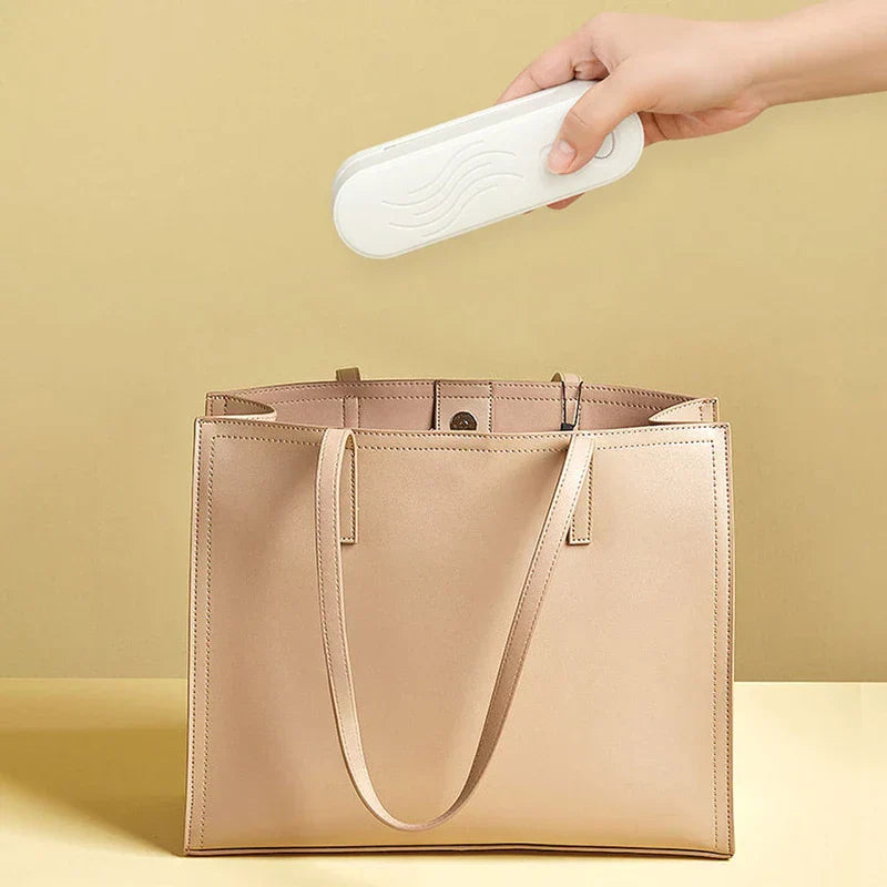 Hand holding a white mini bag heat sealer above an open light tan tote bag, against a solid light yellow background