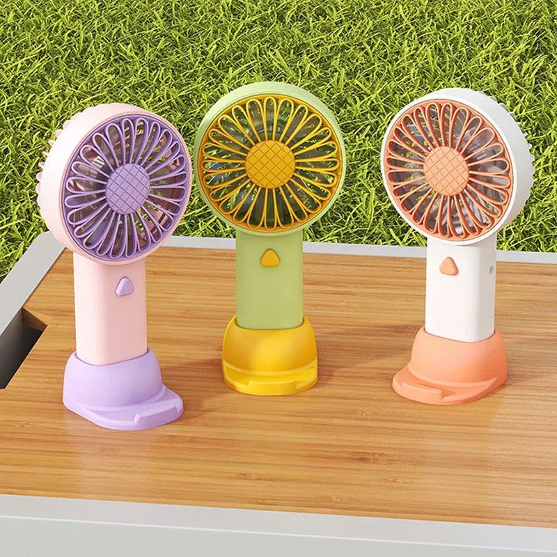 Travel Fan - Three travel fans in pink-purple, green-yellow, and white-orange colorways rest on a wooden surface with grass background