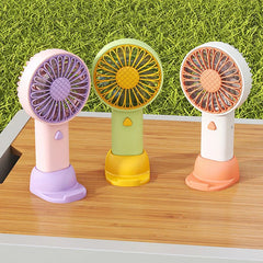 Travel Fan - Three travel fans in pink-purple, green-yellow, and white-orange colorways rest on a wooden surface with grass background
