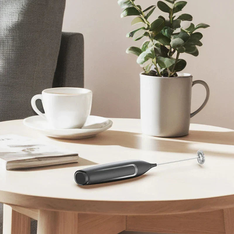 Batidor eléctrico negro para bebidas colocado horizontalmente sobre una mesa redonda de madera junto a una taza de café y una pequeña planta en maceta