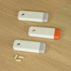 Weekly Pill Organizer - Three weekly pill organizers and three capsule pills on a light wood surface, two with gray ends, one with an orange end