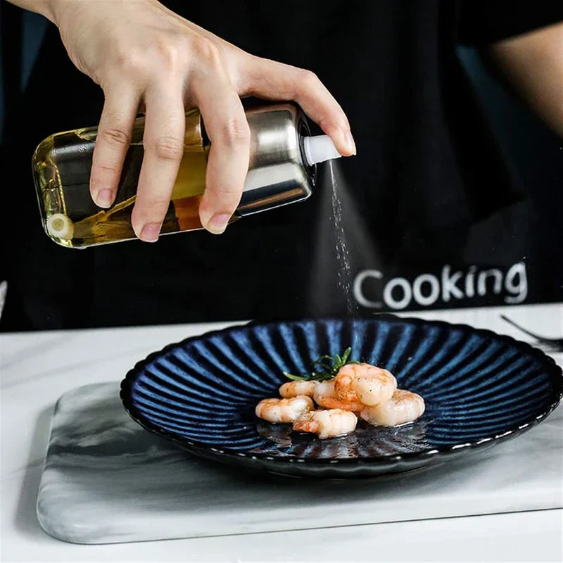 Hand tilts a stainless steel and glass oil sprayer dispenser to mist oil over a plate of cooked shrimp