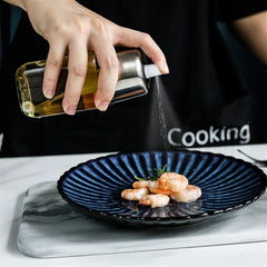 Hand tilts a stainless steel and glass oil sprayer dispenser to mist oil over a plate of cooked shrimp