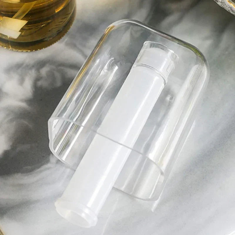 Clear oil sprayer dispenser with internal white tube resting on marbled surface, partial gold-toned jar in corner
