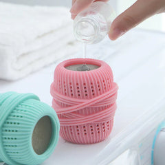 Hand pouring liquid from a glass bottle onto a pink cleaning ball, with a turquoise one beside it on a white surface
