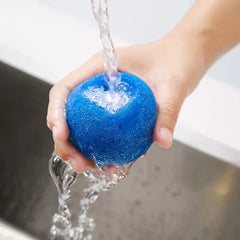 Una mano sosteniendo una bola de lavandería azul porosa bajo agua corriente sobre un fregadero.