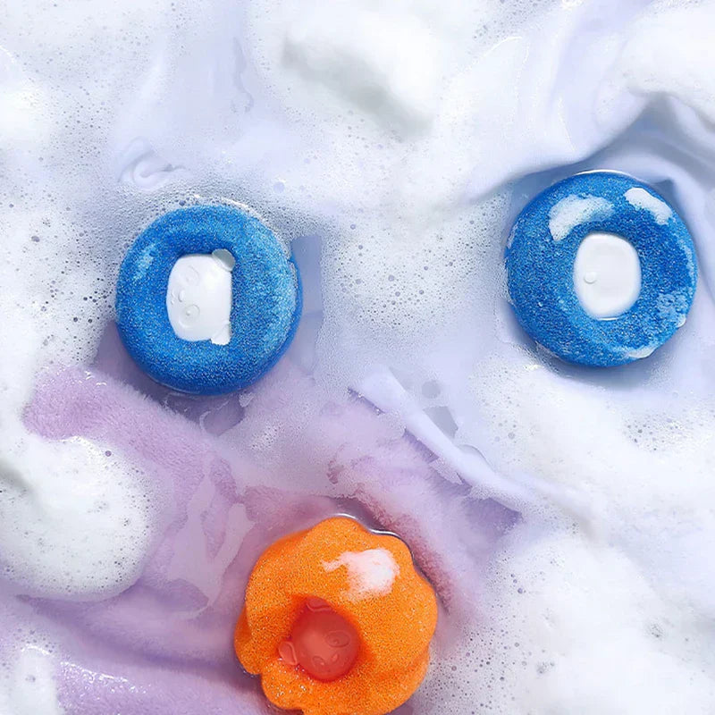bola de lavandería - Dos bolas de lavandería azules y una naranja flotan en agua espumosa de color púrpura claro con burbujas.