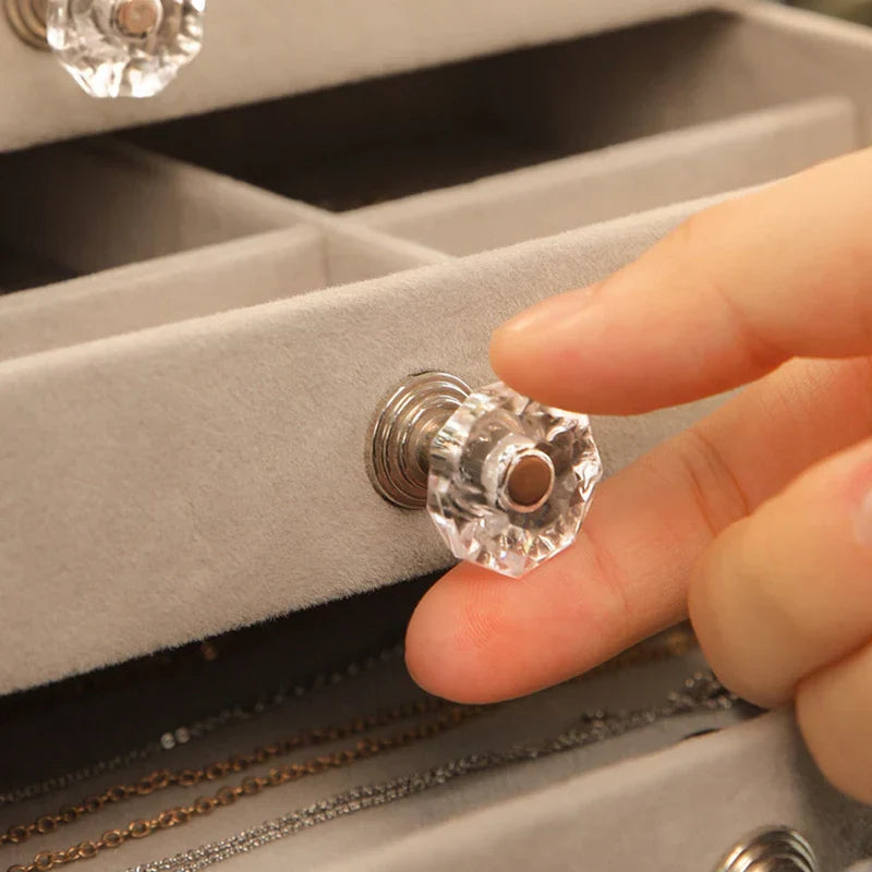 Close-up of a hand holding the clear crystal-style drawer pull of a ring necklace earring display storage box, with a fabric drawer front.