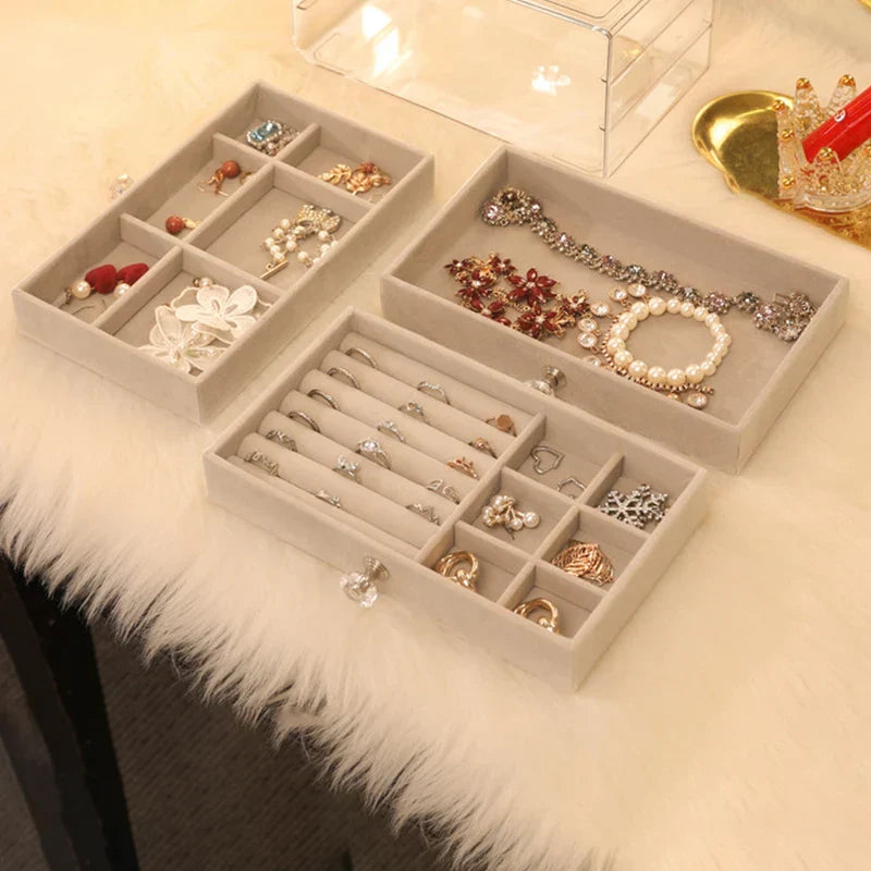 Beige velvet trays holding assorted jewelry sit on a fluffy white surface, with a clear case nearby, featuring a ring necklace earring display storage box.