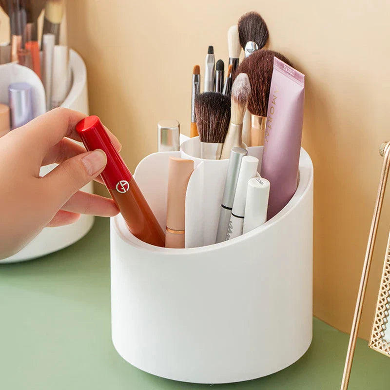 Hand holding red lip gloss above white rotating makeup brush holder filled with brushes and cosmetics on green desk