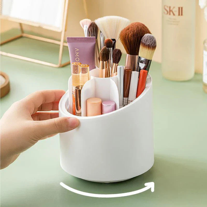 White rotating makeup brush holder filled with makeup brushes and beauty products, hand resting on its side, rotation arrow on green desk