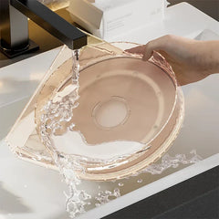 Una mano sostiene un recipiente redondo transparente y poco profundo bajo el agua corriente del grifo, con una escena de tocador de baño relacionada con el gabinete para especias a la vista.