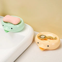 Soap Dish Holder - Two duck-shaped soap dish holders, one green with pink soap on a sink edge, one peach with a gold watch nearby