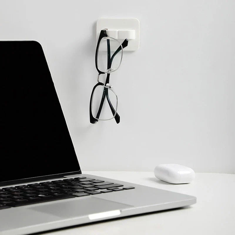 White wall hook hanger holding a pair of black eyeglasses on a wall beside an open laptop and wireless earbud case on a desk