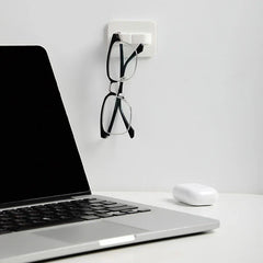White wall hook hanger holding a pair of black eyeglasses on a wall beside an open laptop and wireless earbud case on a desk