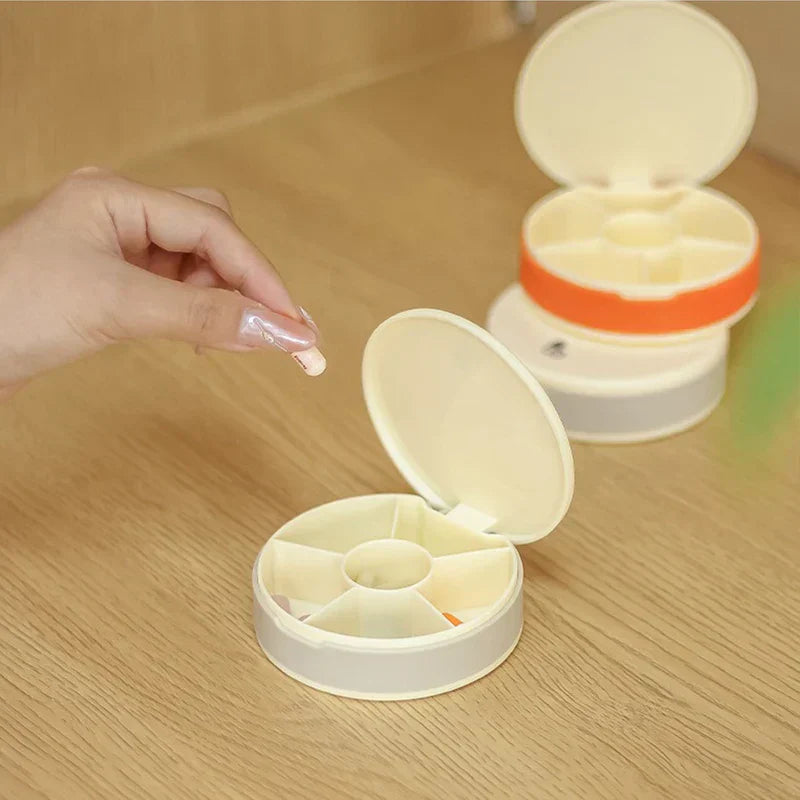 Hand holding a pill near an open gray travel pill organizer box, with an orange-cased one stacked behind on a wood table.