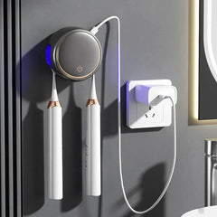 Gray wall-mounted toothbrush sanitizer holder holding two white electric toothbrushes, plugged into a white wall outlet with USB cable