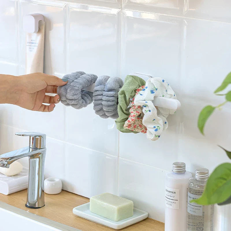 Hand takes a fluffy gray short hair tie from a wall-mounted holder holding multiple hair scrunchies near a bathroom sink faucet.