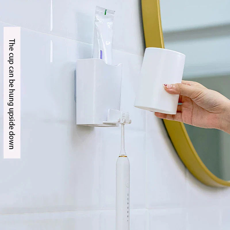 Wall-mounted mouthwash cup holder with a hand holding the removable cup, toothpaste in the slot, and a toothbrush hanging below.