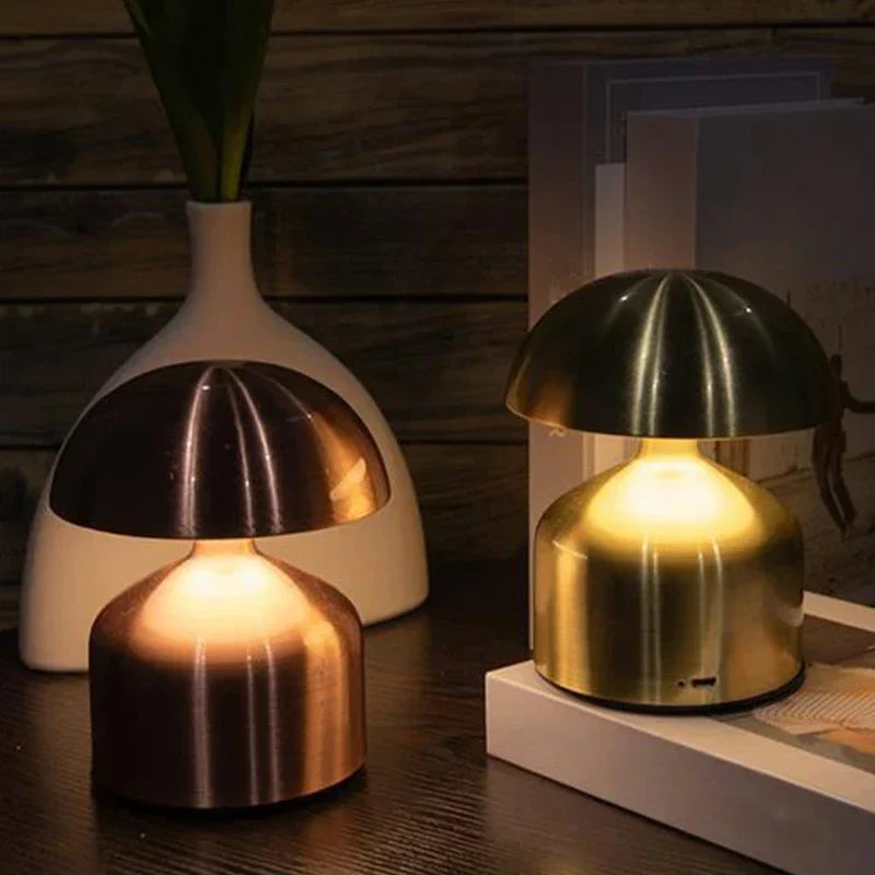 Lámpara de noche LED: dos lámparas de noche LED con forma de hongo en acabado bronce y latón, colocadas sobre una superficie de madera oscura con accesorios decorativos al fondo