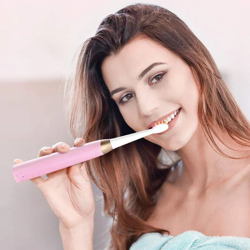 Smiling woman holds a pink electric toothbrush to her teeth while wrapped in a light blue towel