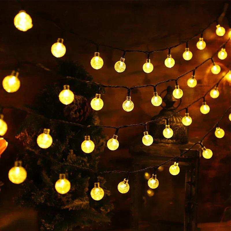 Warm yellow bubble ball string lights drape in multiple rows against a dark backdrop with greenery and a lantern in the background.