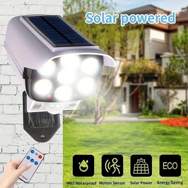 A motion light with solar panel and lit LEDs is mounted on a brick wall, with a remote held below it and a yard in the background.