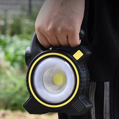 Hand holding black and yellow camping lantern with main light on, outdoors with blurred green background