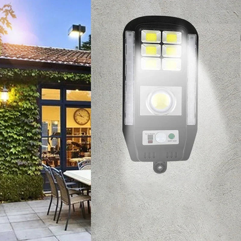 On the right, a solar powered security motion light fixture mounted on a concrete wall; left is a lit patio with outdoor furniture and a house.