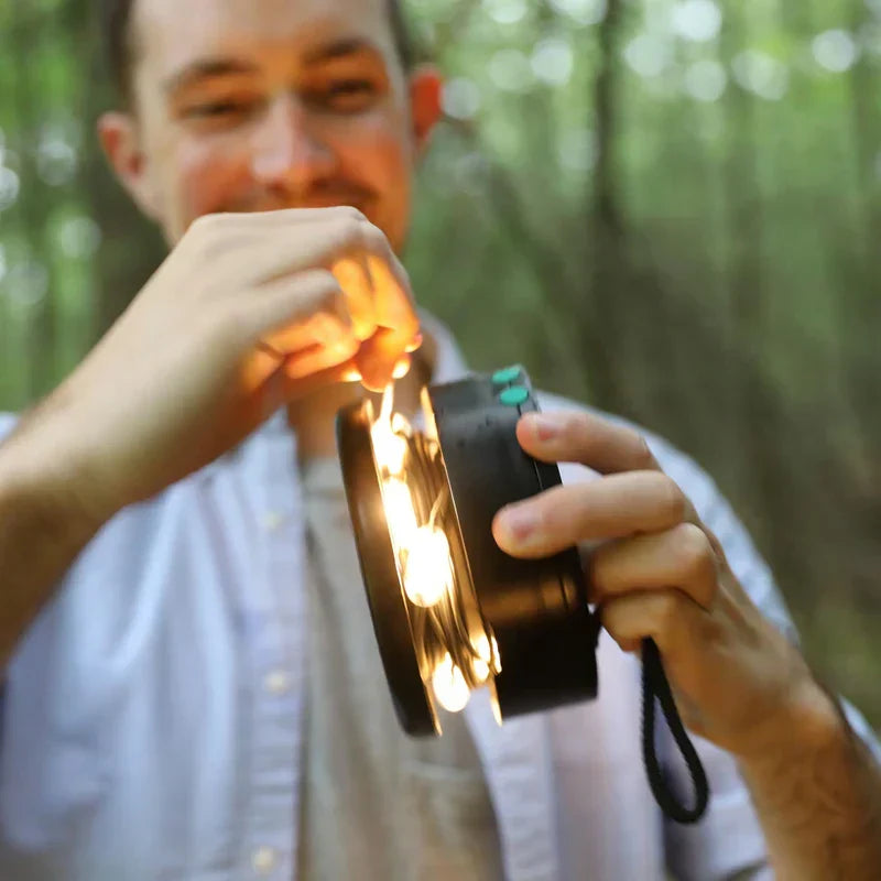 Person holding a lit camping lantern, pulling warm string lights from its side outdoors in woods