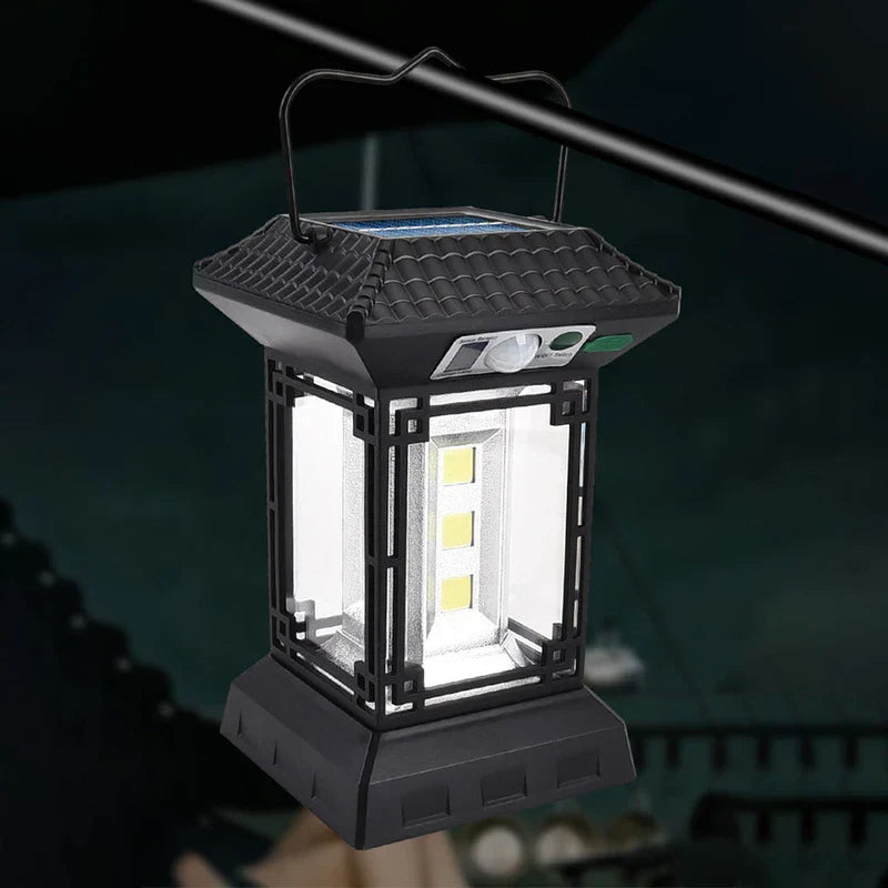 luz de campamento - Vista frontal angular de linterna negra estilo pagoda iluminada colgando de un poste metálico, mostrando botones de control, sensor de movimiento y paneles LED encendidos contra un fondo de campamento oscuro desenfocado