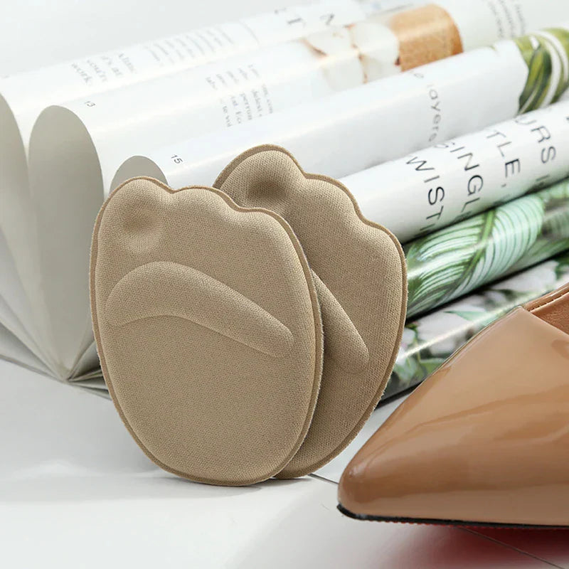 almohadillas de esponja para el antepié - Dos almohadillas para el antepié de color beige descansando sobre una superficie blanca, junto a la punta puntiaguda de un zapato color nude con libros enrollados al fondo