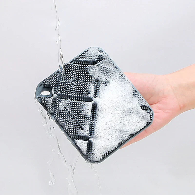cepillo de ducha para el cuerpo - Una mano sostiene una almohadilla de cerdas cuadrada gris mientras el agua enjuaga la espuma