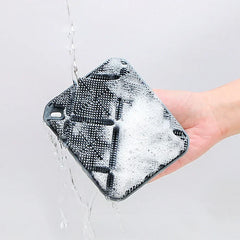 cepillo de ducha para el cuerpo - Una mano sostiene una almohadilla de cerdas cuadrada gris mientras el agua enjuaga la espuma