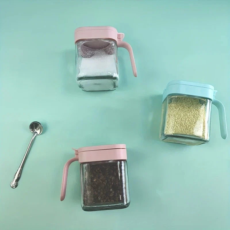 Frasco de vidrio para especias - Tres recipientes cuadrados llenos con tapas rosa y azul claro colocados sobre superficie menta, con cuchara metálica pequeña a la izquierda