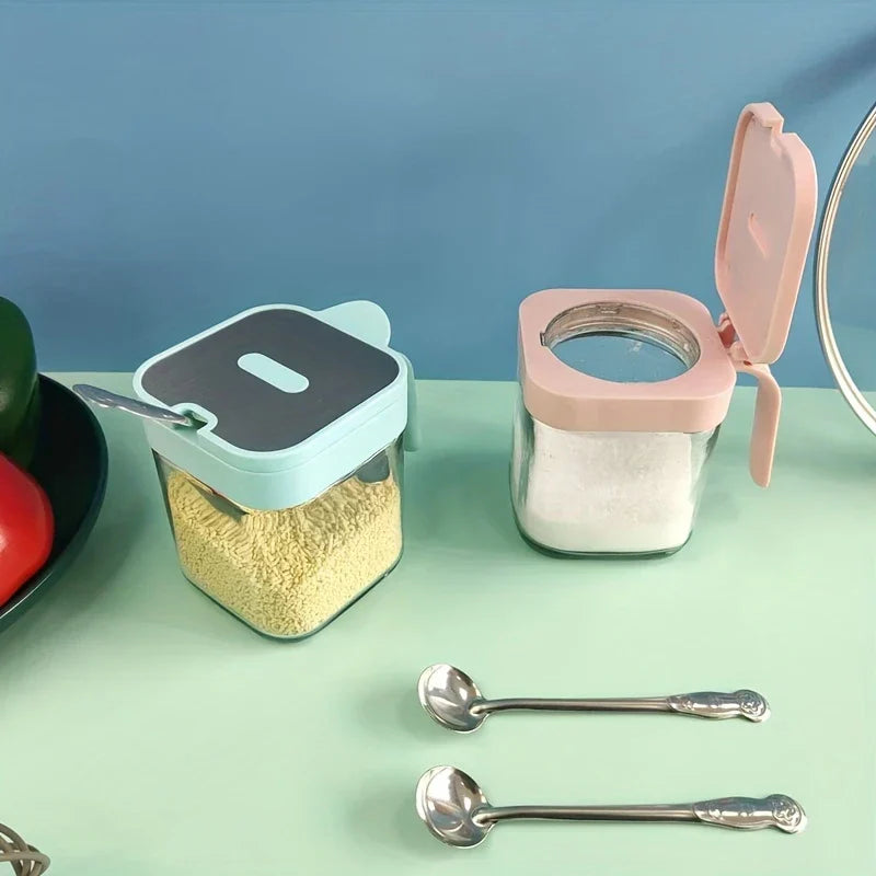 Frasco de vidrio para especias - Dos recipientes cuadrados llenos sobre encimera menta junto a pimientos, con dos cucharas metálicas pequeñas al frente y una tapa abierta