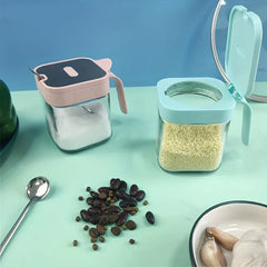 Glass Spice Jar - Two glass spice jars on a light green countertop, one pink-lidded holding salt with a spoon, one blue-lidded open holding granular seasoning, with small items nearby.