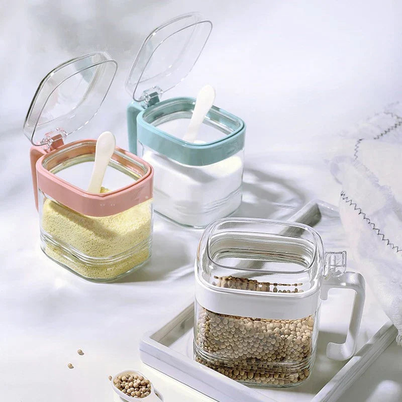 Glass Spice Jar - Three glass spice jars with open lids: two on cloth holding granular seasonings with spoons, one on a tray holding seeds, spoon with seeds nearby.