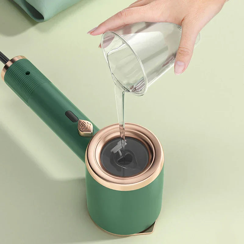 Prensa de prendas - mano vertiendo agua de un vaso transparente al depósito de agua de un vaporizador de prendas portátil verde y rosa dorado