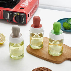 escurridor de vidrios - Tres frascos con pincel para aceite con tapas de colores, uno sobre una encimera, dos sobre una pequeña tabla de cortar de madera junto a una hornilla portátil, limón y plato con brócoli