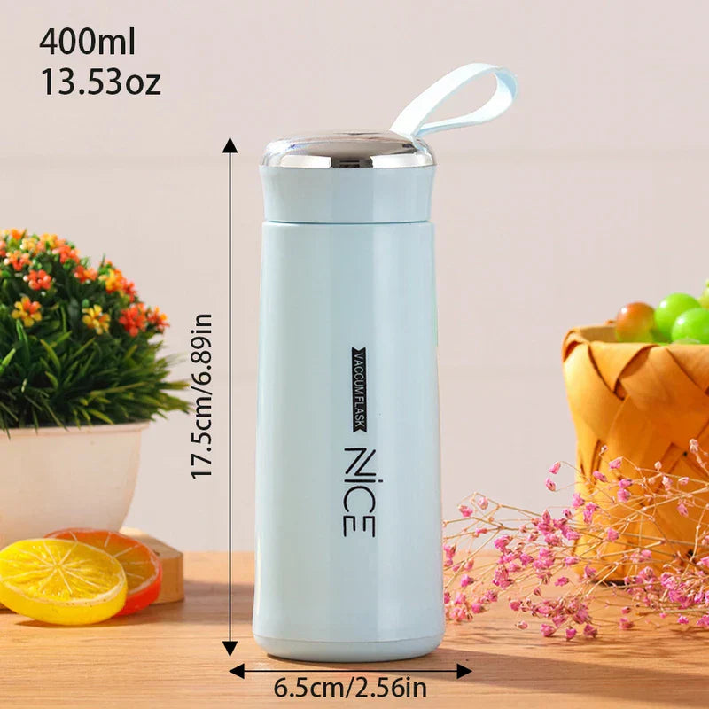 Light blue plastic tumbler cup with carry strap sits on a wooden table, with dimensions labeled. Small decor items sit nearby.