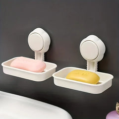 Two white wall-mounted suction cup soap dish holder units, one holding pink soap and one holding yellow soap, against a dark wall