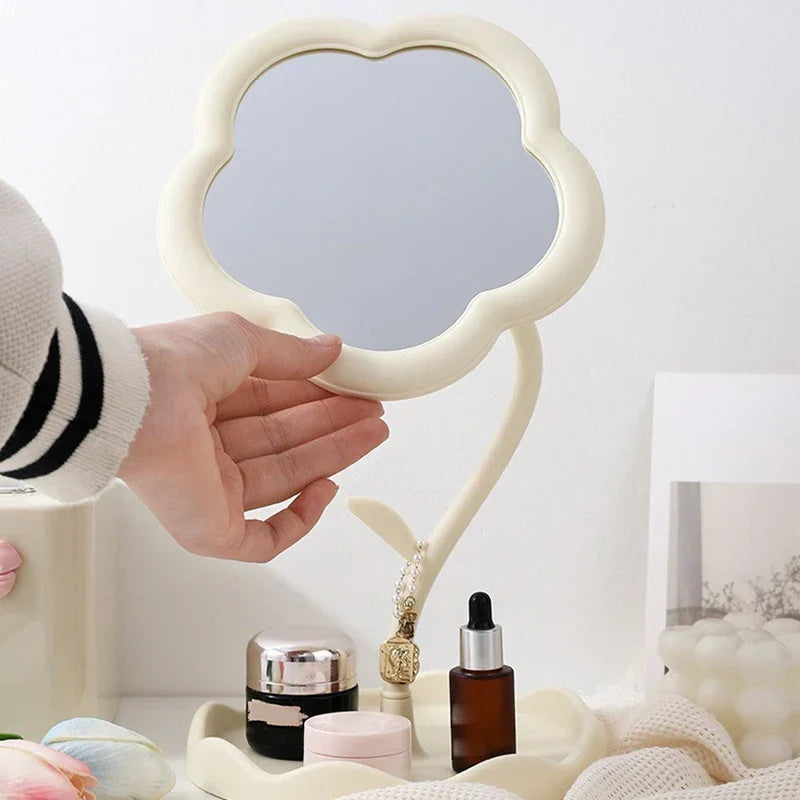 Makeup Mirror - Cream flower-shaped desktop mirror with a hand touching its frame, base tray holding small skincare bottles, surrounded by desk decor