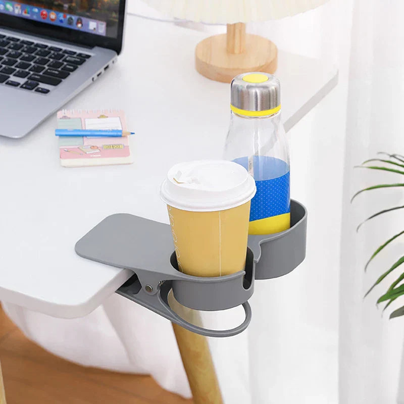 Soporte con pinza para bebidas de mesa - Portabebidas gris con clip sujeto al borde de un escritorio blanco, sosteniendo un vaso de café desechable para llevar y una botella de agua transparente junto a una computadora portátil abierta y un pequeño bloc de notas